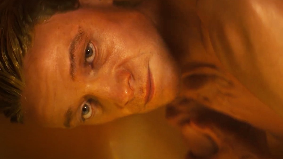 A sideways close-up shows an orange-lit man with short facial stubble, light-colored eyes, and a faint smile while looking toward the camera, with another blurred figure or body part in the background. The warm, hazy behind-the-scenes framing matches the post’s teaser for DC’s “Clayface,” which is noted as coming to theaters on October 23.