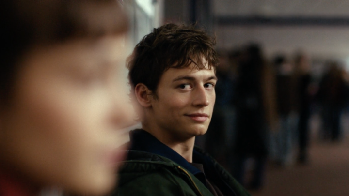 A young man with tousled dark hair glances over his shoulder with a subtle, knowing smile at someone out of focus in the foreground, likely in a crowded indoor setting that suggests a school or social gathering, fitting the tense, youthful drama promised by "Storm" arriving on Prime Video.