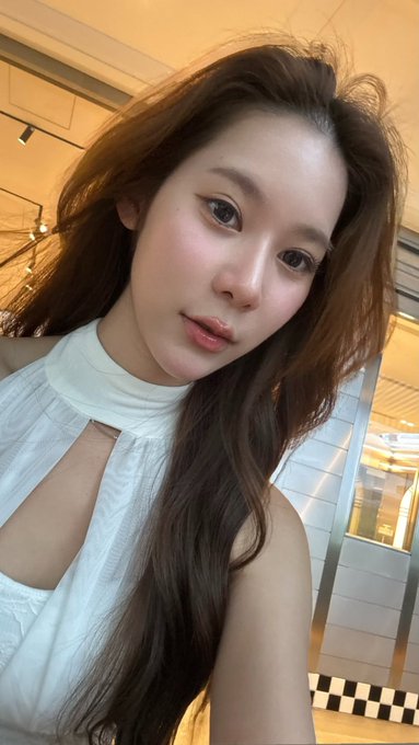 A close-up selfie of a young woman with long brown hair wearing a white halter top, taken indoors.