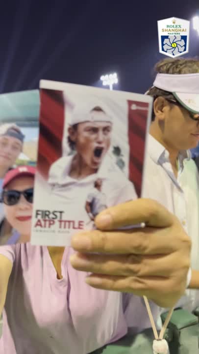 A tennis fan holds up a card celebrating "FIRST ATP TITLE" featuring a player image, with the Rolex Shanghai Masters logo visible in the background.