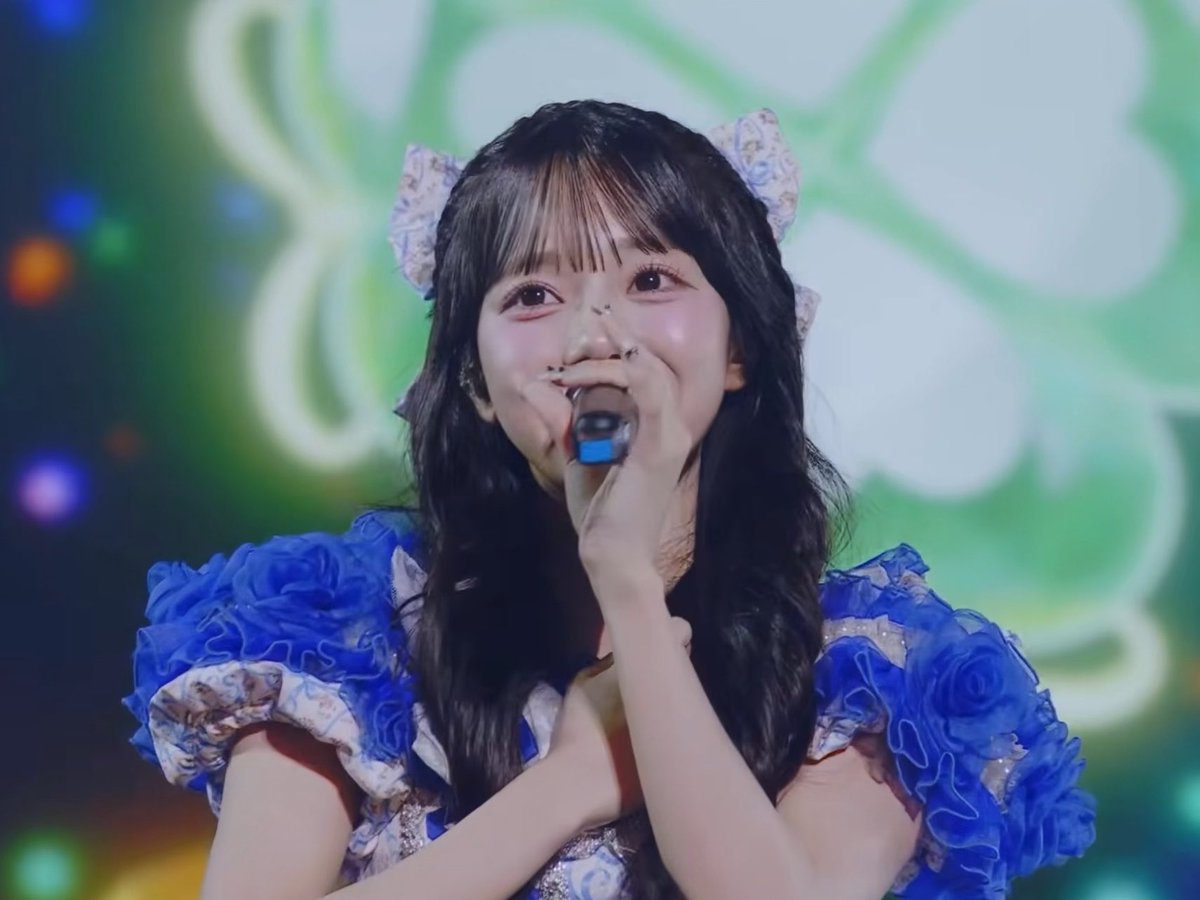 A young woman with long dark hair, wearing a blue floral ruffled dress and hair bows, sings into a microphone on stage with a green lit background.
