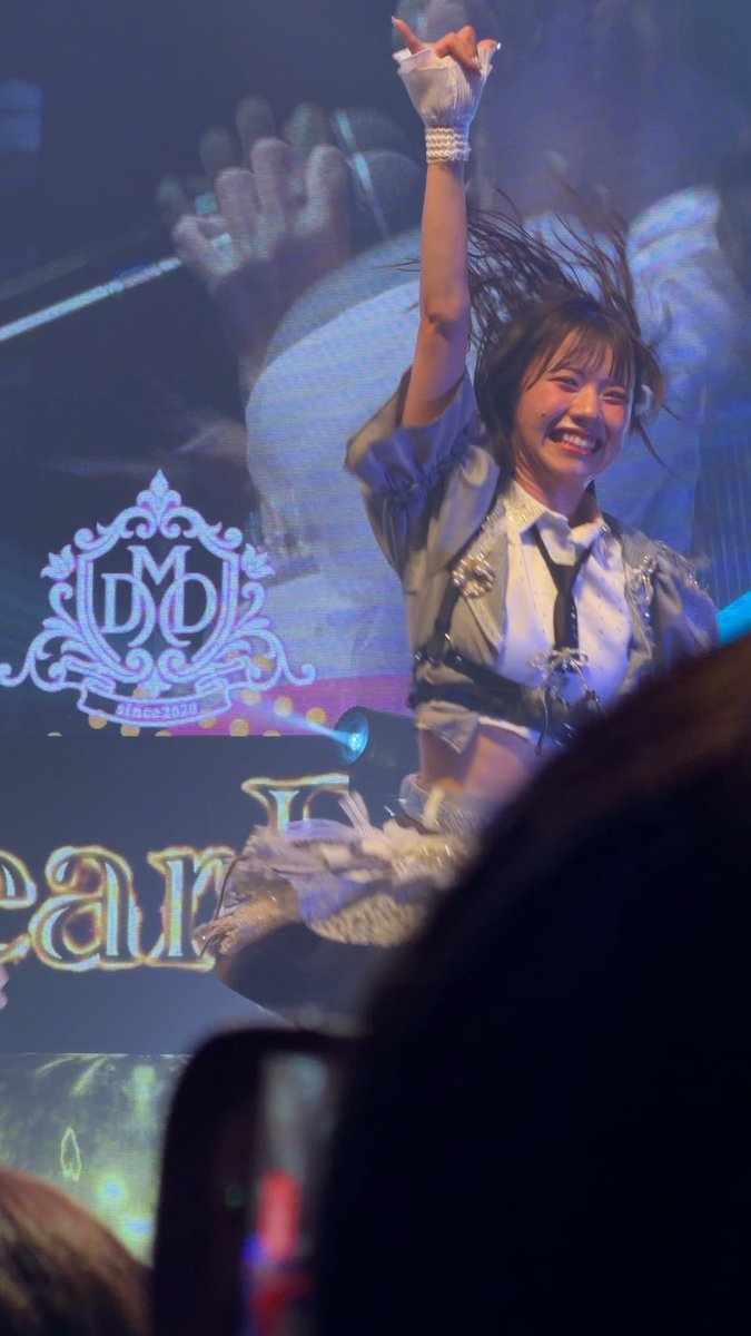 A performer on stage smiling with her arm raised, wearing a white and grey outfit with a lace glove, in front of a screen displaying a 'since 2020' crest logo and partial text reading 'ean'.