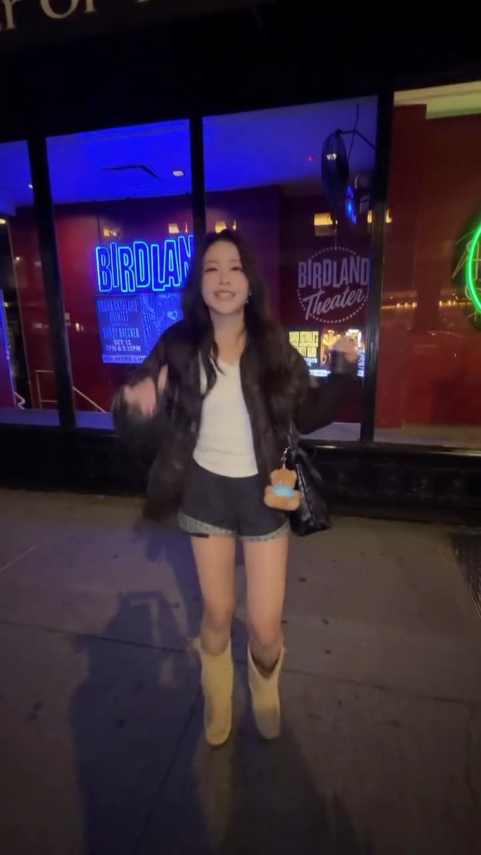 A young woman stands smiling on a sidewalk at night in front of the neon-lit "Birdland Theater" marquee, wearing a fur jacket, white top, denim shorts, and tan boots while carrying a black bag.
