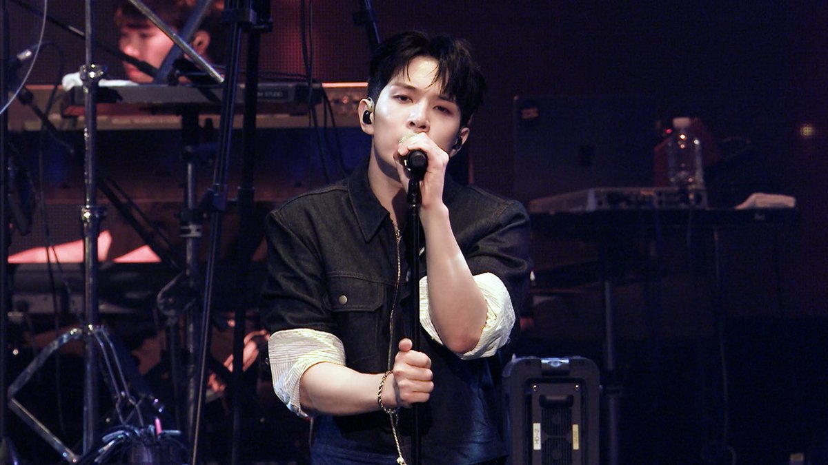 A performer with dark hair, wearing a black denim jacket and in-ear monitors, sings into a microphone on stage, illuminated by concert lighting with musical equipment visible in the background, capturing a moment from N.Flying's live performance of "Still."