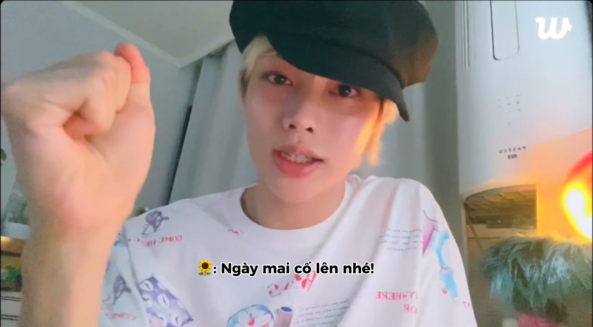 A selfie-style video (Weverse platform) of a person wearing a patterned white t-shirt and black cap, with Vietnamese subtitle text '🌻: Ngày mai cố lên nhé!' meaning 'Tomorrow, do your best!'