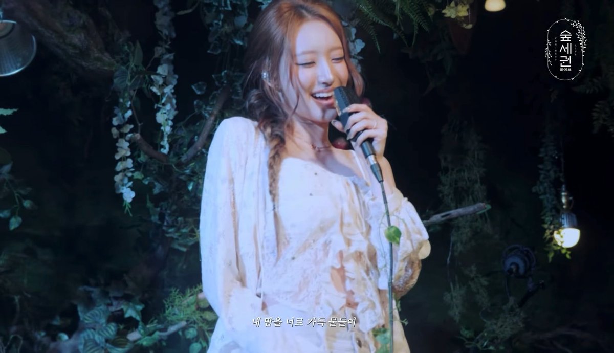 A woman with reddish-brown hair in a braided style smiles while singing into a microphone; she wears a white dress against a dark, foliage-decorated background, with Korean subtitles below and a logo reading '金세련 HOUSE' in the top right.