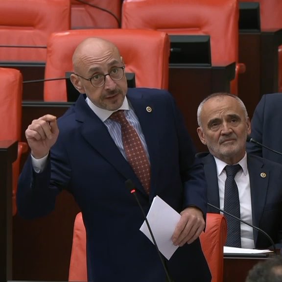 A bald man wearing glasses and a blue suit with a red patterned tie speaks at a podium in what appears to be a parliamentary chamber with red seats, while another man in a suit sits behind him.