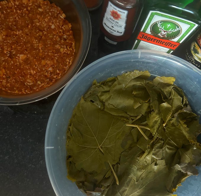 A top-down view of food preparation showing a bowl of grape leaves (likely for dolmas), a bowl of chili flakes, a bottle of "Indian Balt Sauce", and a bottle of Jägermeister in the background.