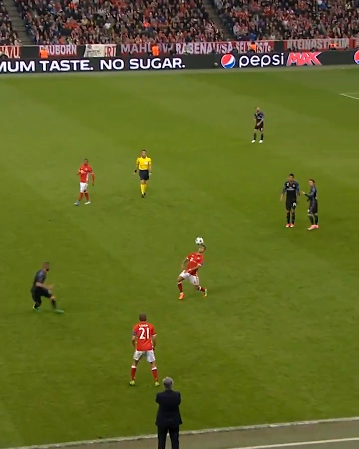 A still from a soccer match showing a player in a red uniform controlling the ball while players in dark blue and a referee in yellow look on.