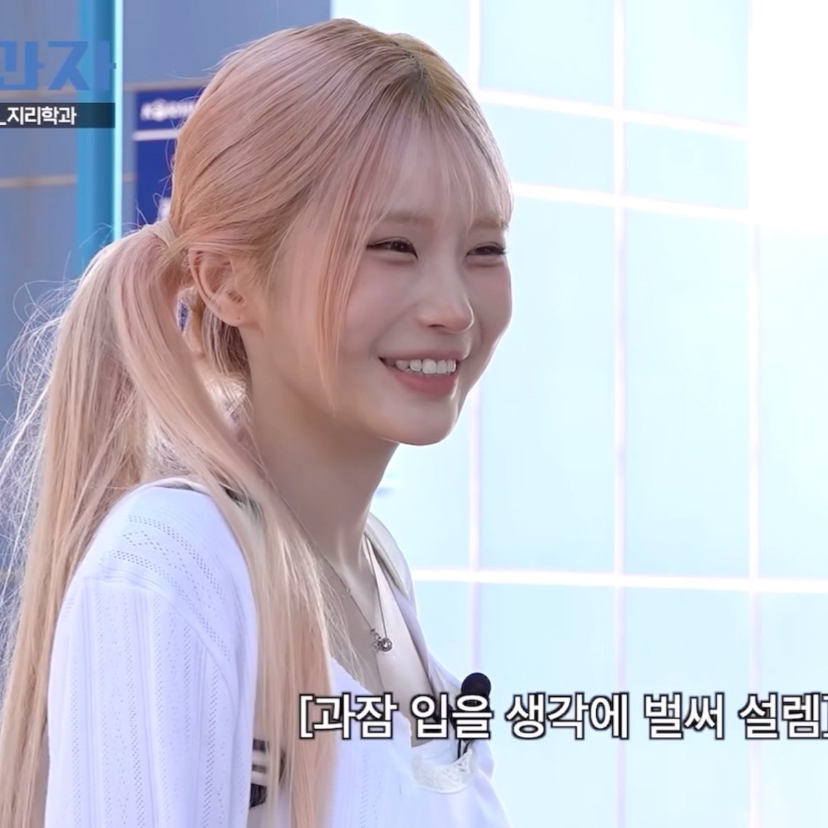 A Korean broadcast screenshot shows a smiling young woman with long pinkish hair wearing a white top, with Korean subtitles reading "[과장 임을 생각에 벌써 설렘!" at the bottom.