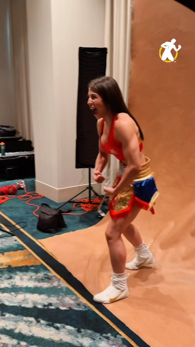A muscular woman in a red crop top and blue/gold Muay Thai shorts celebrating with clenched fists in what appears to be an indoor training or event venue, with a small yellow figure sticker overlay.