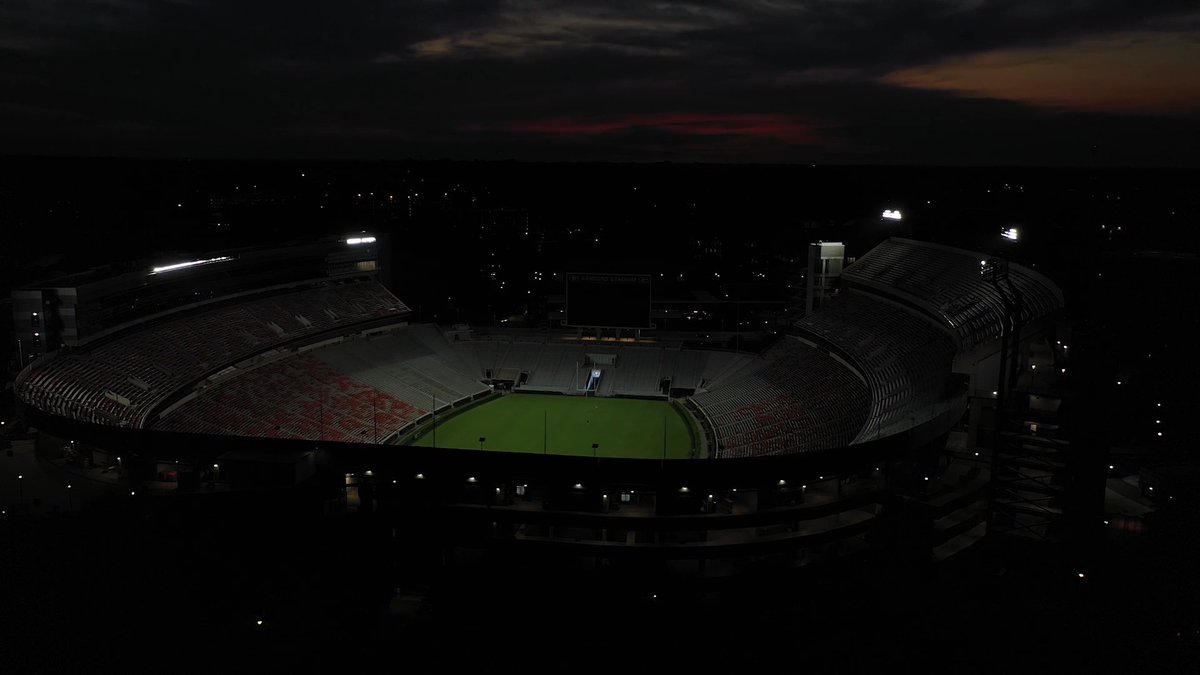 A darkened Sanford Stadium on the University of Georgia campus is shown at dusk, illuminated only by ambient lights highlighting the empty field and stands. The image connects to the post about a partnership with Hunker Spirits, a Georgia-based distillery founded by UGA alumni, reflecting the shared Georgia roots celebrated by the new collaboration.