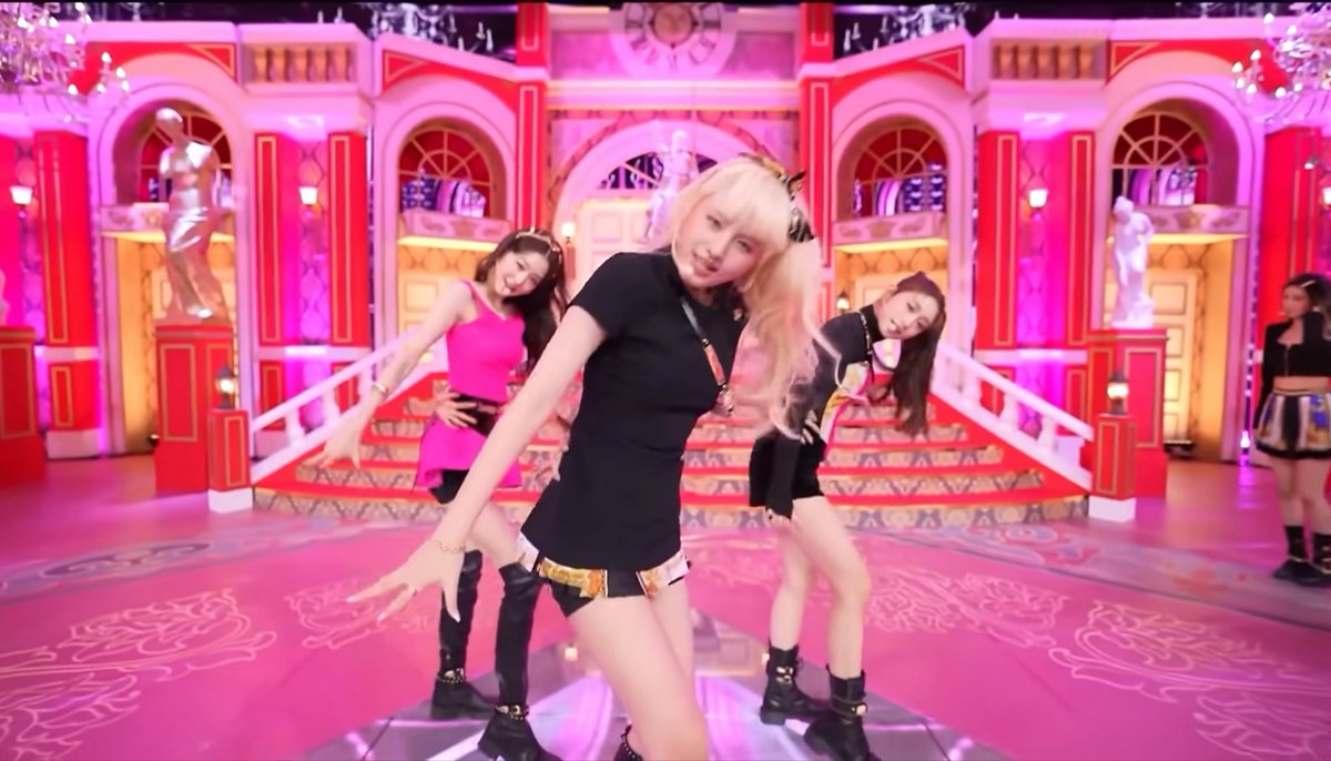 Members of the K-pop group Blackpink performing a dance routine on an elaborate pink-themed set resembling a grand palace, with the blonde member Rosé in the foreground wearing a black top and shorts.