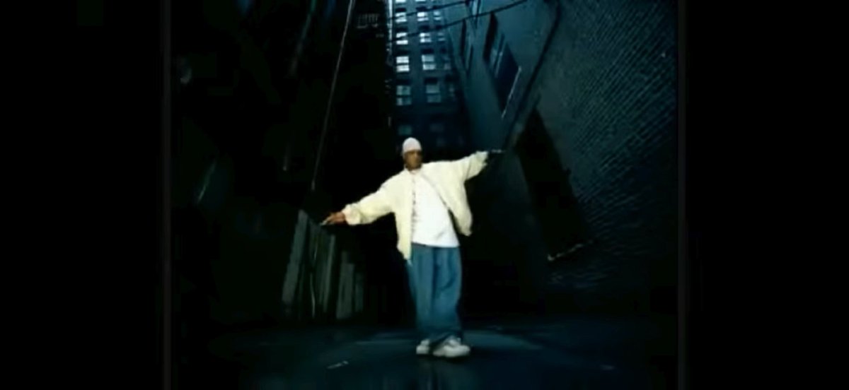 A performer in a white outfit and beanie stands center stage under dramatic lighting, arms outstretched against a dark industrial backdrop in what appears to be a music video scene.