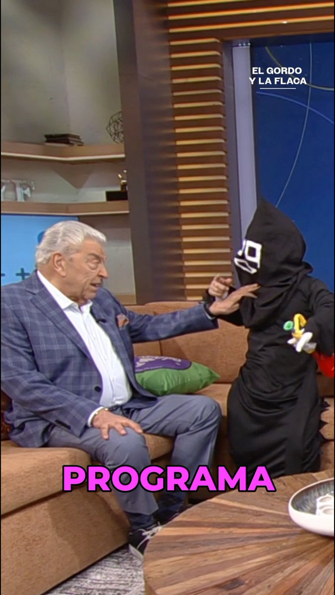 A man in a blue plaid blazer sits on a brown couch on a TV set, gesturing toward a person dressed in a black hooded costume with a mask; the image has the label 'EL GORDO Y LA FLACA' and the text 'PROGRAMA' in pink.