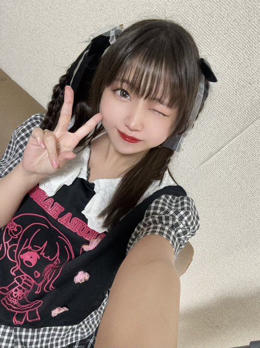 A woman taking a selfie, winking and making a peace sign, wearing a black and white checkered top with a black bib featuring a pink character design.