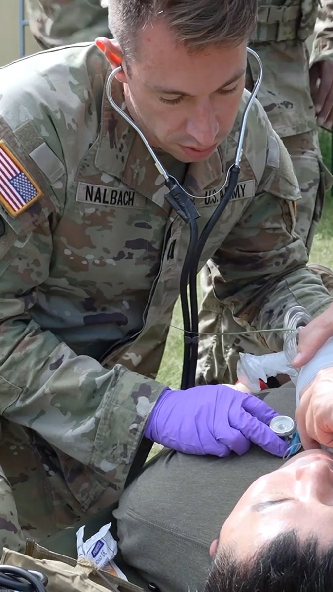 A U.S. Army soldier wearing purple medical gloves uses a stethoscope to examine a patient lying on the ground during a field medical training exercise; the soldier's name tape reads 'NALBACH'.