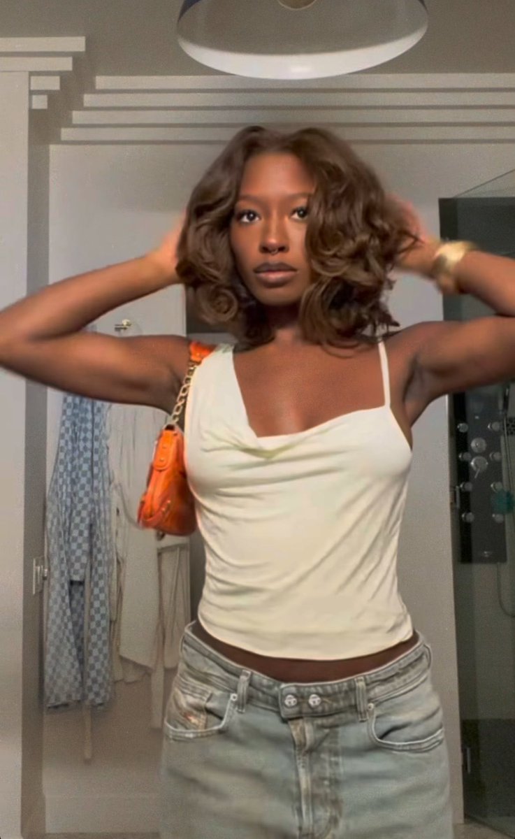 A mirror selfie of a young woman with wavy brown hair wearing a light-colored tank top, denim skirt, and carrying an orange purse.