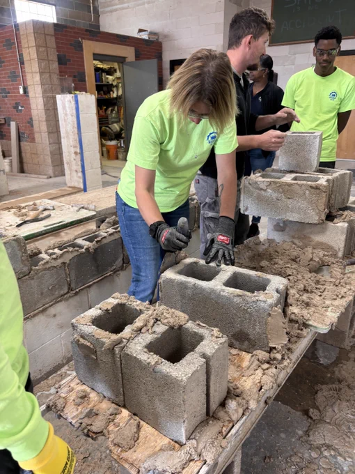 Individuals in bright green shirts are engaged in a hands-on masonry task, working with cement blocks on a sturdy table in a construction workshop setting. The environment includes tools and building materials.