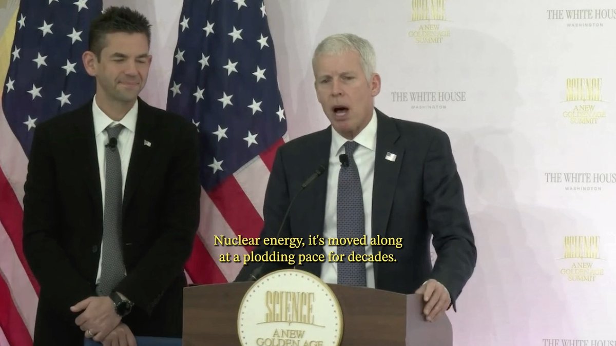 Secretary Wright speaks at the podium during the New Golden Age Summit at the White House, with a subtitle reading "Nuclear energy, it's moved along at a plodding pace for decades," while another man in a suit stands beside him in front of American flags, illustrating his call for a nuclear renaissance.