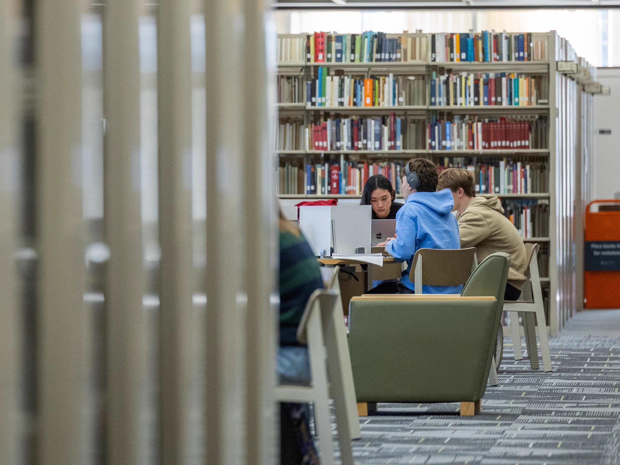 A group of Tulane students sits at a table in a library, working on laptops. They are surrounded by tall bookshelves filled with various books. The setting appears quiet and studious.