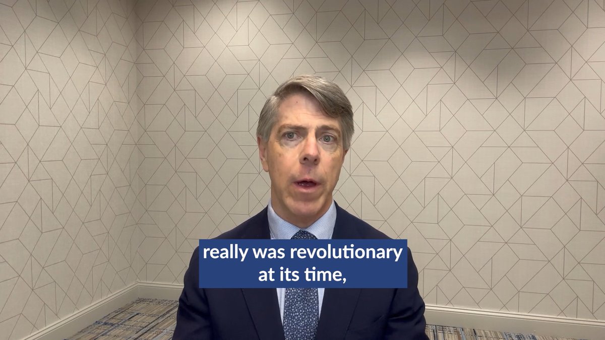 A man in a dark suit and patterned tie stands in a corner room with geometric patterned wallpaper, with subtitle text reading "really was revolutionary at its time,"