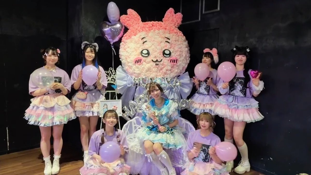 A group of seven performers in purple and blue idol outfits pose together with pink balloons and a large pink rabbit piñata.