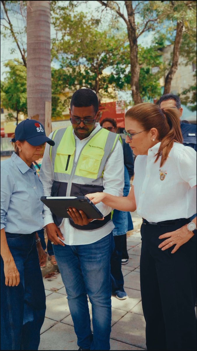 Three people standing outdoors looking at a tablet device together: a woman in an 'SB' cap, a man in a yellow safety vest labeled 'Ing. Alisson Bonnelly', and a woman in a white shirt with an emblem.