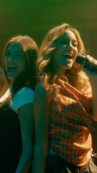 Two smiling young women with long hair, one holding a microphone, appearing to sing or perform under stage lighting.