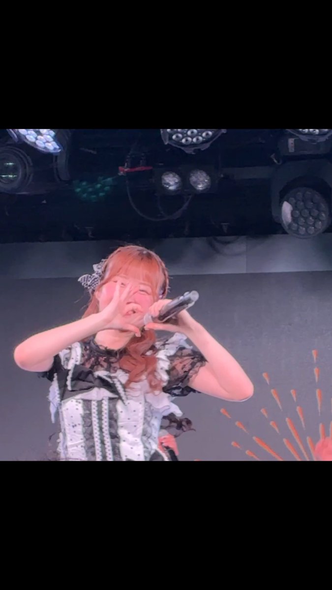 A young performer with orange hair, wearing a black and white ruffled dress and a bow, singing into a microphone on a stage with stage lights visible in the background.