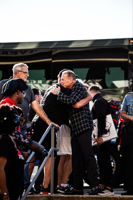 Two older men embrace and speak into a microphone in a crowded outdoor setting, with a cameraman and other personnel visible nearby.