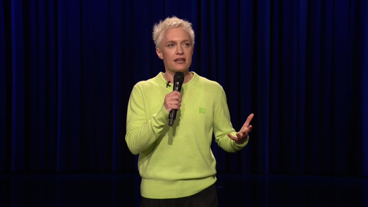 A person with short blonde hair wearing a green sweater holds a microphone while performing stand-up comedy on stage against a dark blue curtain backdrop, illustrating a clip from a comedy segment. The caption aligns with the post's mention of Emma Willmann's joke about her landlord "Gary" on the Tonight Show.