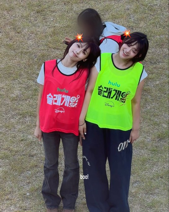 Two young women smile and pose close together on a grassy field while wearing red and neon green sports bibs branded with 'hulu' and '슬라게임' (Disney's 'Slam Game'), with 'boo!' watermarked on the image.