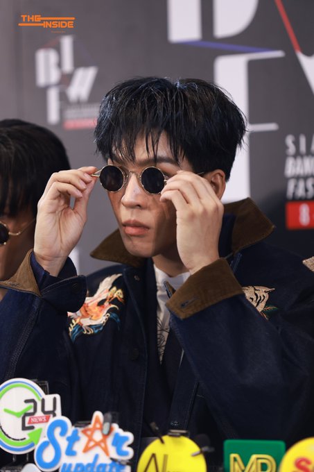 A news photo from GMM 24 (The Inside Star Update) showing a man with dark hair adjusting round sunglasses while wearing a denim jacket with embroidered designs on the collar.