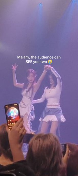 A fan's video screenshot from a concert showing two female performers on stage holding hands up while wearing white outfits, with audience members' phones visible in the foreground. Overlaid text reads 'Ma'am, the audience can SEE you two 😭'.