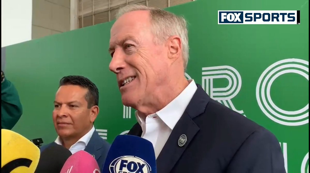 A man in a suit being interviewed by Fox Sports microphones in front of a green backdrop, with another man standing beside him.