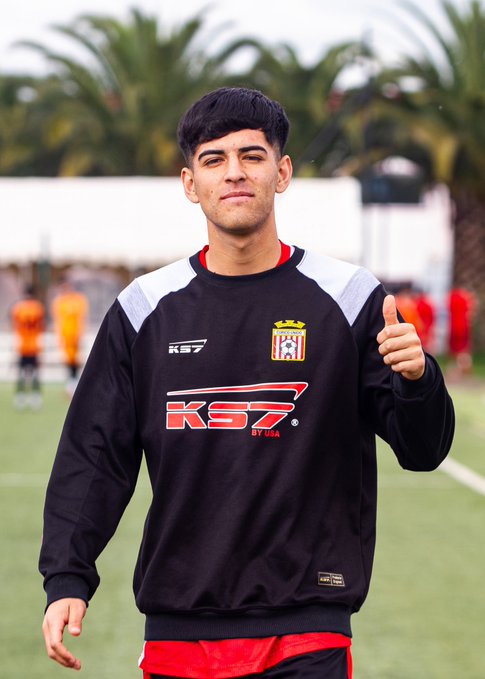 A young male soccer player with dark hair stands on a field giving a thumbs-up, wearing a black, white, and red sweatshirt with 'KS7' branding and a team crest.