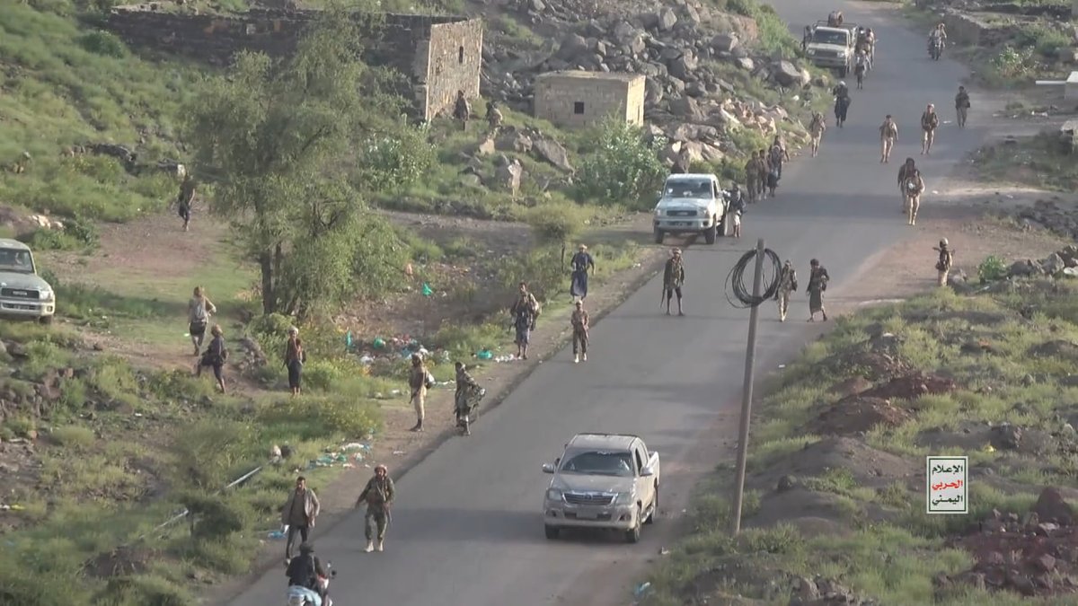 An aerial view of a road in a rocky, rural area with armed soldiers patrolling and several pickup trucks, with Arabic text in the bottom right corner.