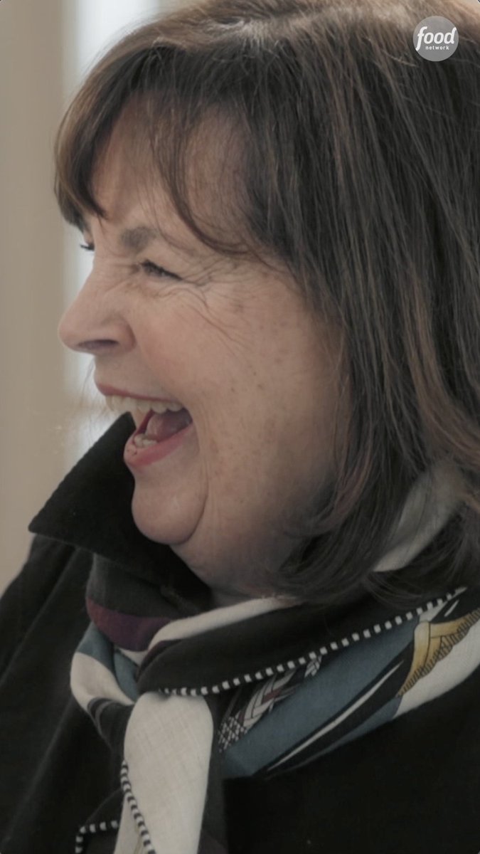A close-up profile shot of Ina Garten laughing, wearing a dark jacket and a patterned silk scarf, with a Food Network logo visible in the corner.