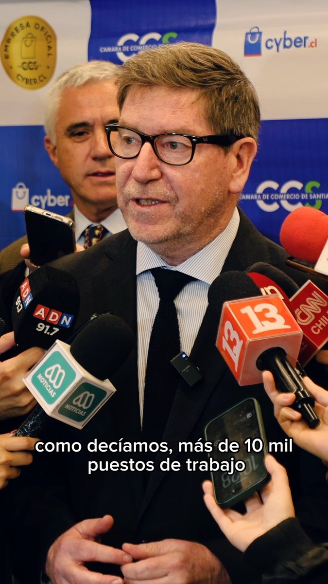 A bespectacled man in a suit speaks to multiple media microphones including ADN, Canal 13, CNN Chile, and Mega, with a subtitle reading 'como decíamos, más de 10 mil puestos de trabajo'.