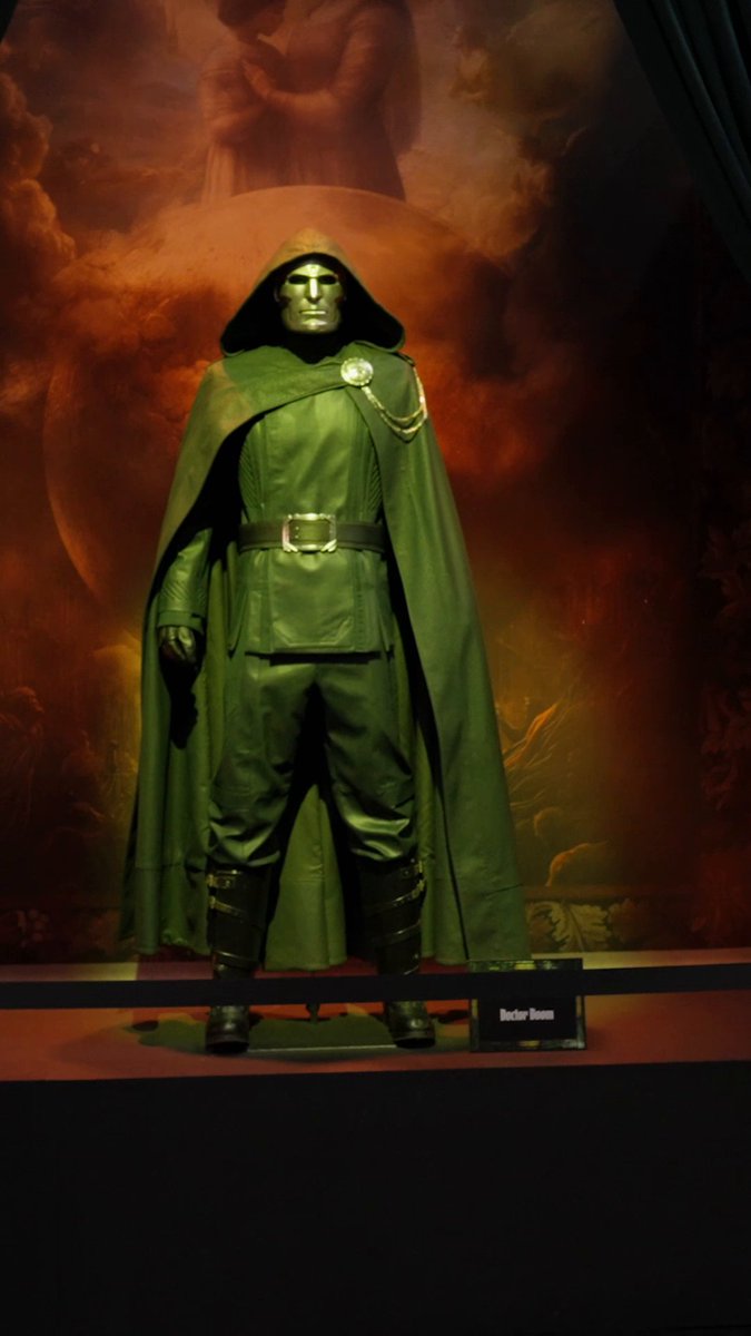 A display of the Doctor Doom costume—a green hooded cloak, metallic mask, and utility belt—standing on a platform with a 'Doctor Doom' nameplate, against a dramatic red backdrop.