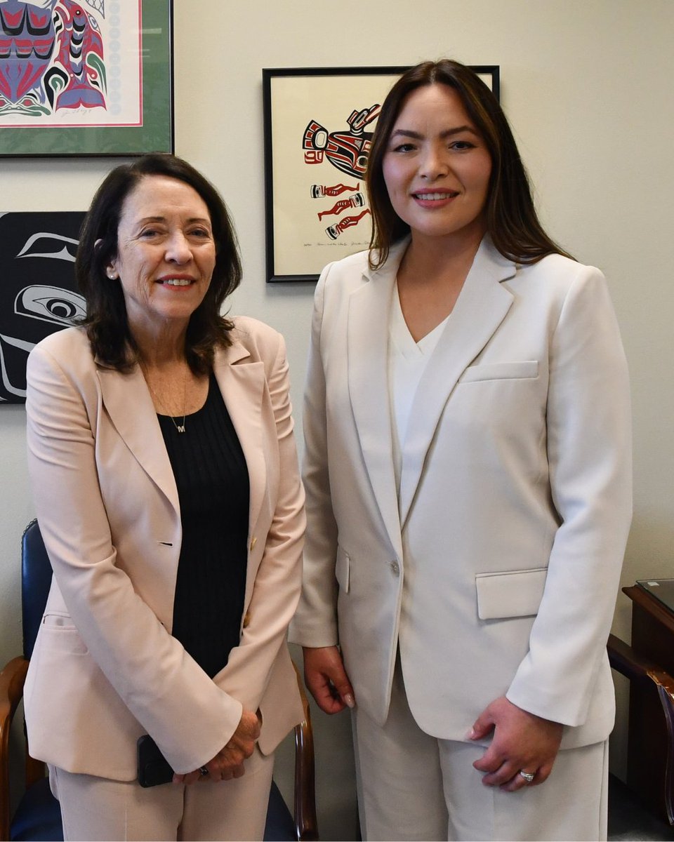 Two women in light-colored suits pose together indoors in front of framed Indigenous-style artwork. No prominent text is visible.