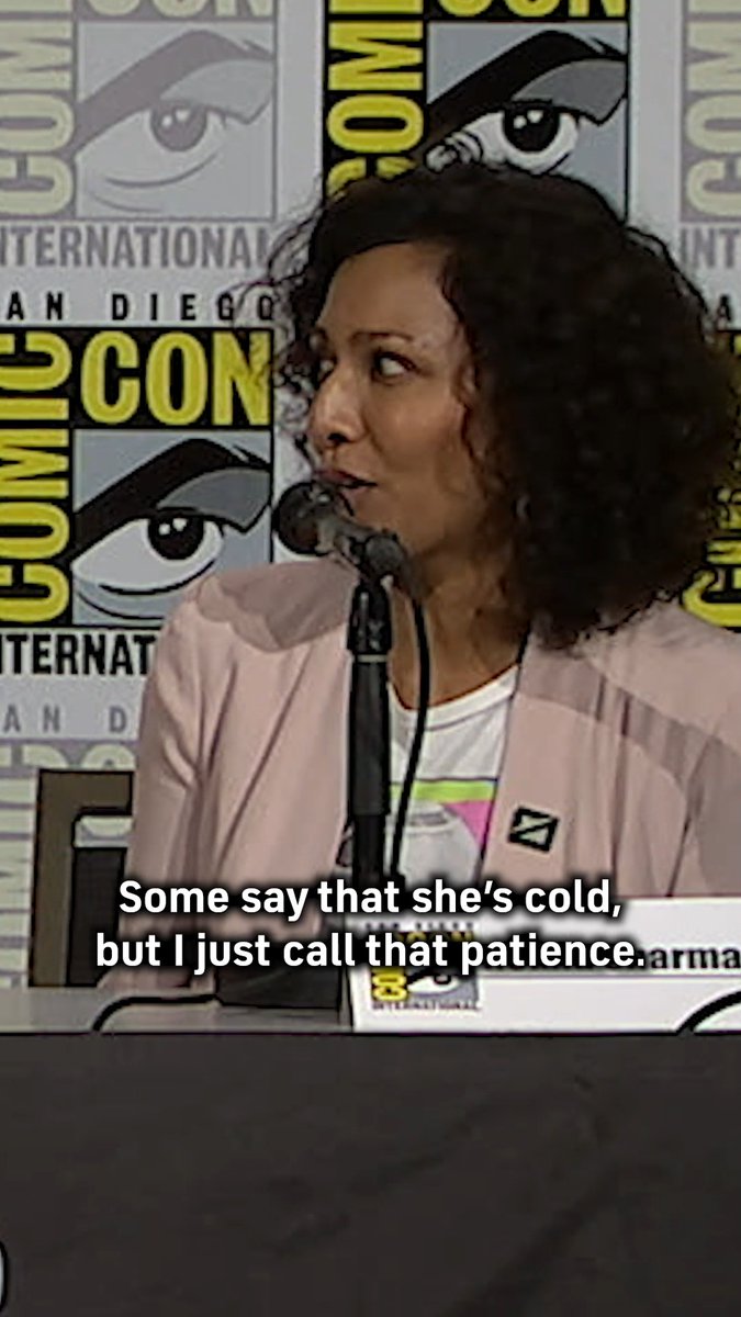 A woman with curly hair speaks into a microphone at a San Diego Comic-Con panel, wearing a name badge partially reading "arma," with overlaid text reading "Some say that she's cold, but I just call that patience." The image illustrates the anecdote about Rekha Sharma discussing her character Fathom's "patient" nature at the #SWZeroCompany panel.