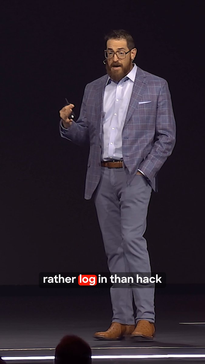 A man in a plaid suit and glasses giving a presentation on a dark stage, holding a clicker, with a caption overlay reading 'rather log in than hack' with 'log' highlighted.