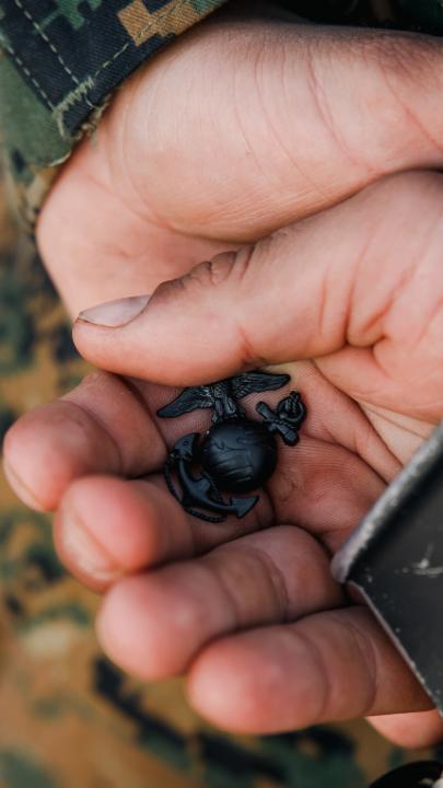 A close-up of a hand holding the black Eagle, Globe, and Anchor insignia of the United States Marine Corps, the iconic emblem signifying a Marine's completion of recruit training and earned status. The image visually represents the rigorous achievement referenced in the post, emphasizing that Marine Corps judge advocates must meet the same demanding standards as all other Marines before serving as lawyers.