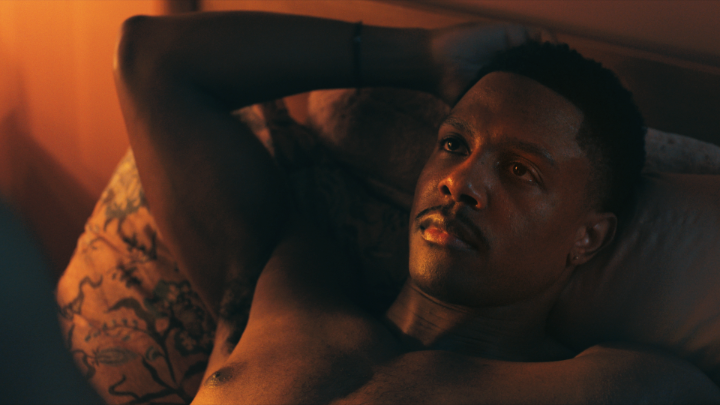 A shirtless man with tattoos on his right arm lies on a patterned pillow in dimly lit, warm orange and blue lighting, resting his hands behind his head and gazing upward with a contemplative expression.