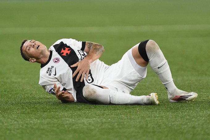 A Vasco da Gama soccer player, wearing a white and black jersey featuring the distinctive red cross club crest, lies on the grass field clutching his abdomen in apparent pain. The image illustrates the concerning moment of an injury during the match, prompting teammate Bruno Duarte to publicly reassure fans about the player's condition following the scare.