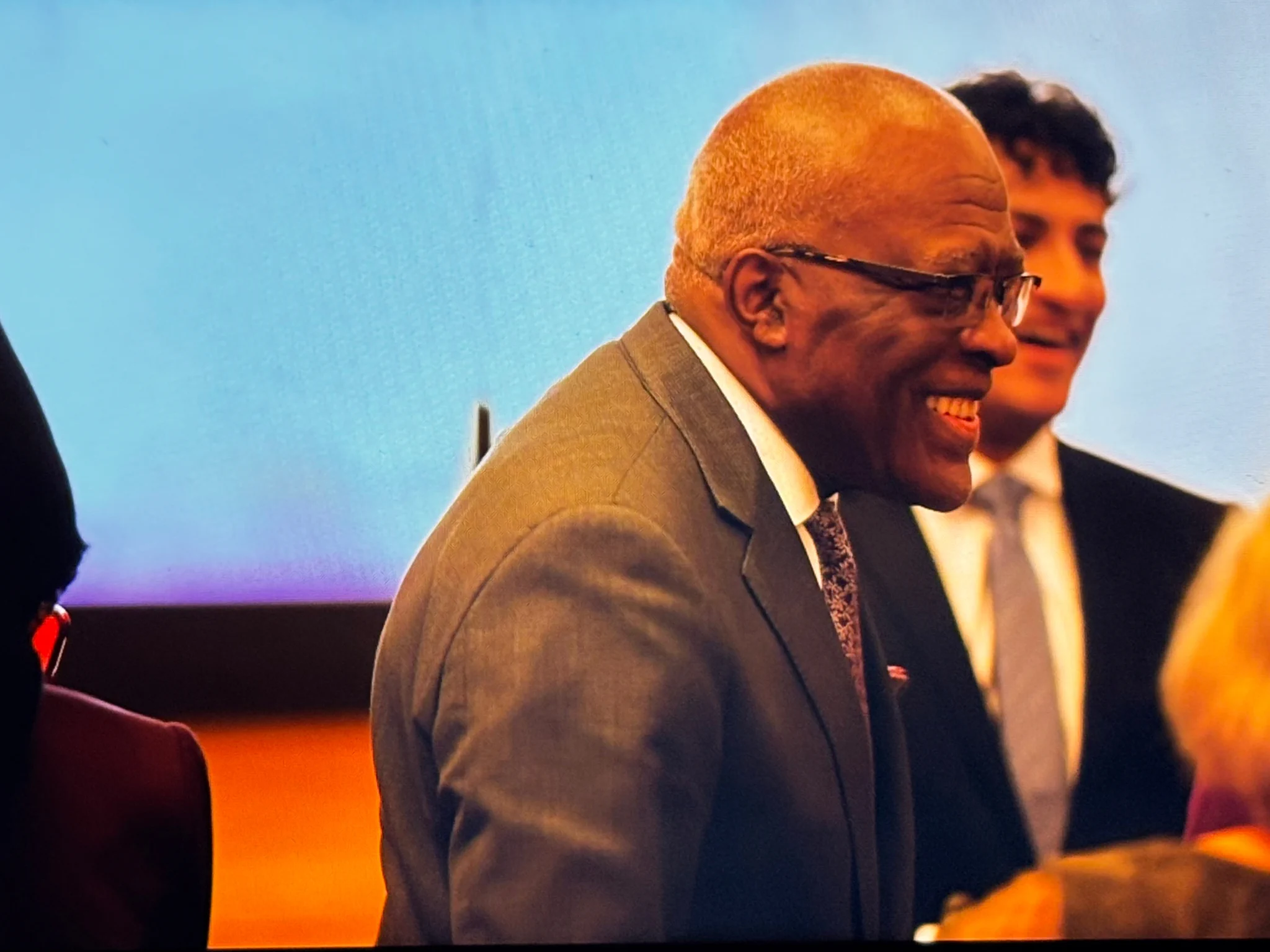 University of Washington President Robert J. Jones in a grey suit and glasses smiles widely, looking right. Behind him, a blurred man with dark hair also smiles. Light blue background.