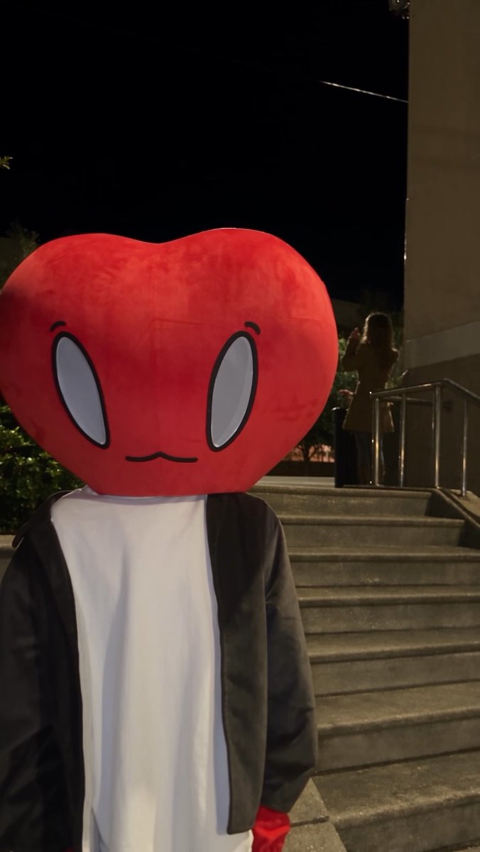A person in a black jacket and white shirt stands on outdoor steps wearing a large, plush red heart head with cartoon eyes and a wavy mouth.