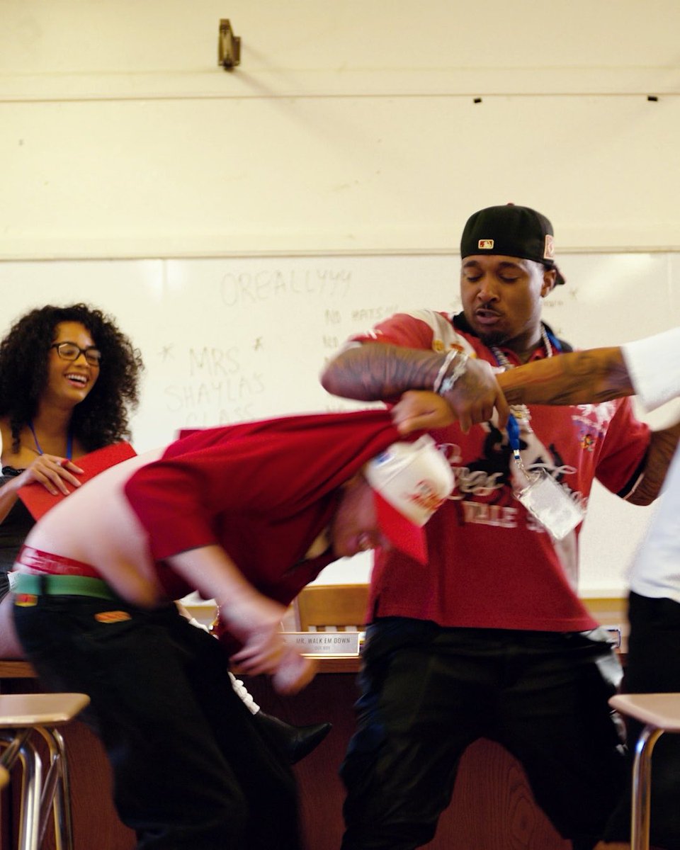 Two men in red shirts engage in a physical altercation in what appears to be a classroom setting, with one man appearing to pull at the other's shirt; a smiling woman with curly hair and glasses watches from the left near a whiteboard with text including 'CREALLY!!' and 'MRS SHAYLAS CLASS'.